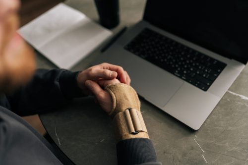 Close-up top view of male hands wearing wrist brace, massaging painful wrist while working remotely from home office, surrounded by laptop and notebook. Concept of struggles of remote work