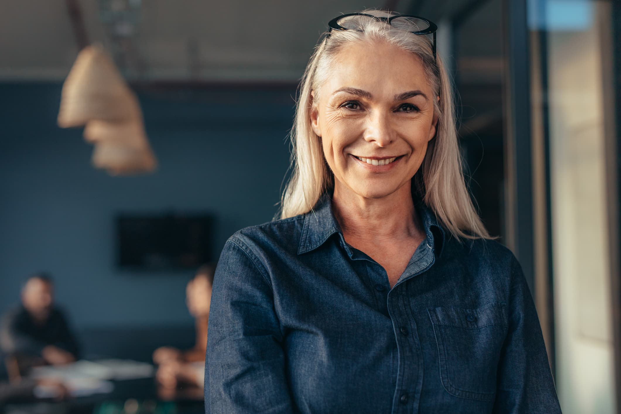Close up of senior business woman standing in office looking at camera and smiling. Mature female in office with team in background.