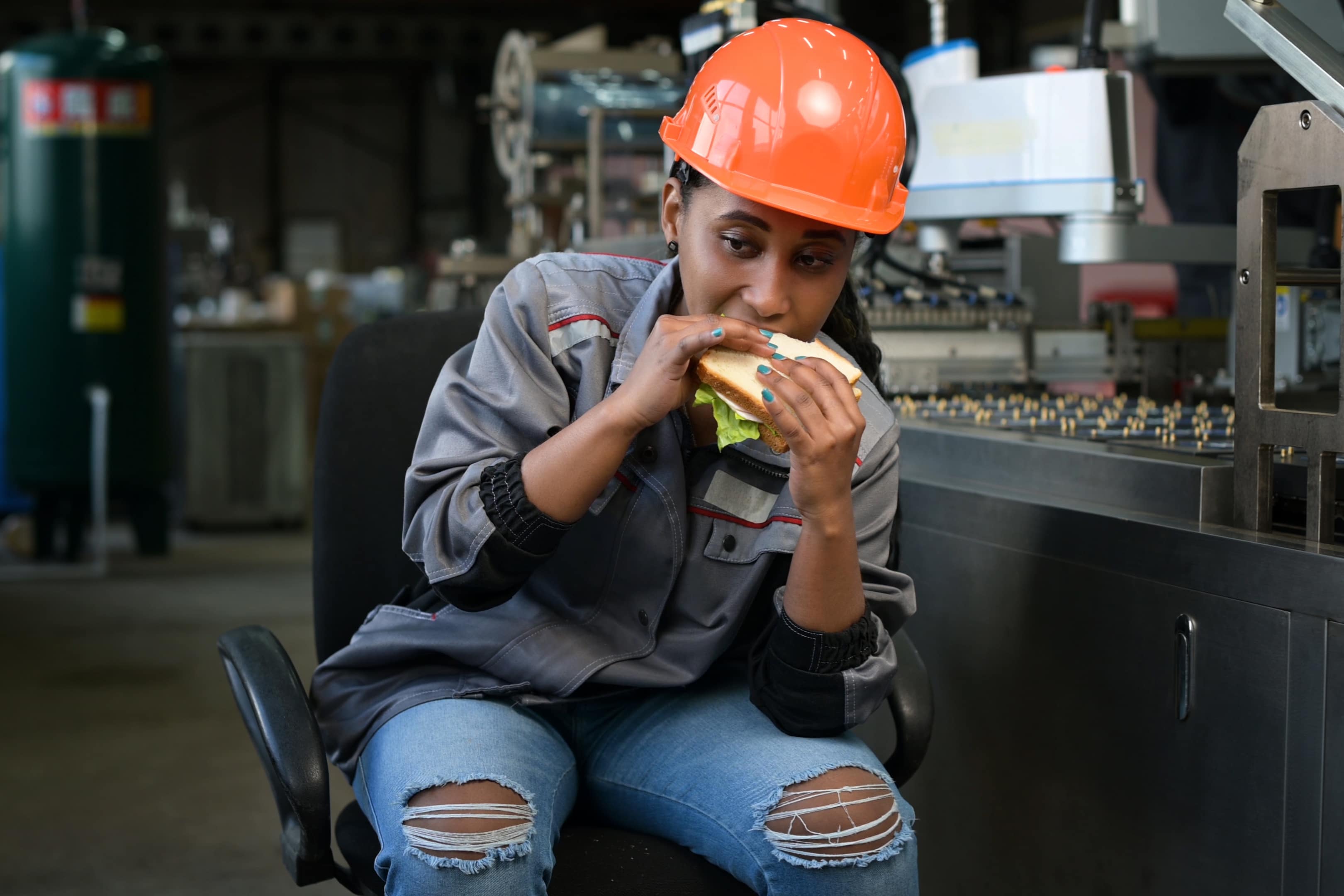 A young black woman in an orange helmet eats a sandwich in a production facility, next to a conveyor belt. Production worker taking a lunch break at the workplace