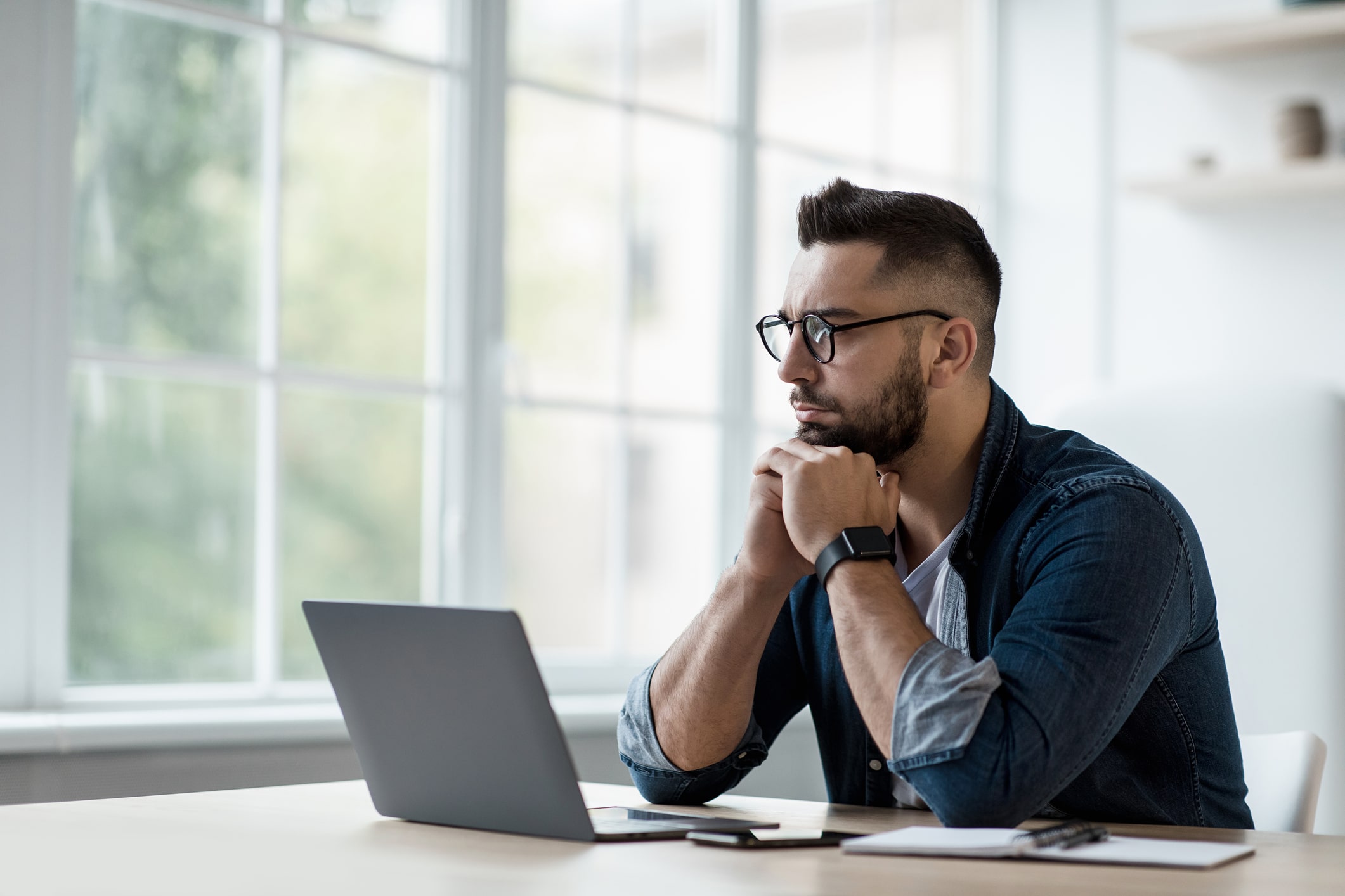 Serious young attractive man in glasses, thinks, watch online lesson, in interior with laptop and phone