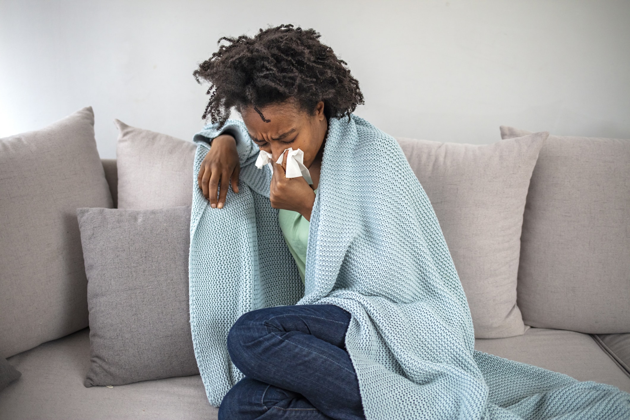 Sick young african woman feeling cold covered with blanket sit on sofa, ill black girl shivering freezing warming at home wrapped with plaid, no central heating problem, fever temperature flu concept