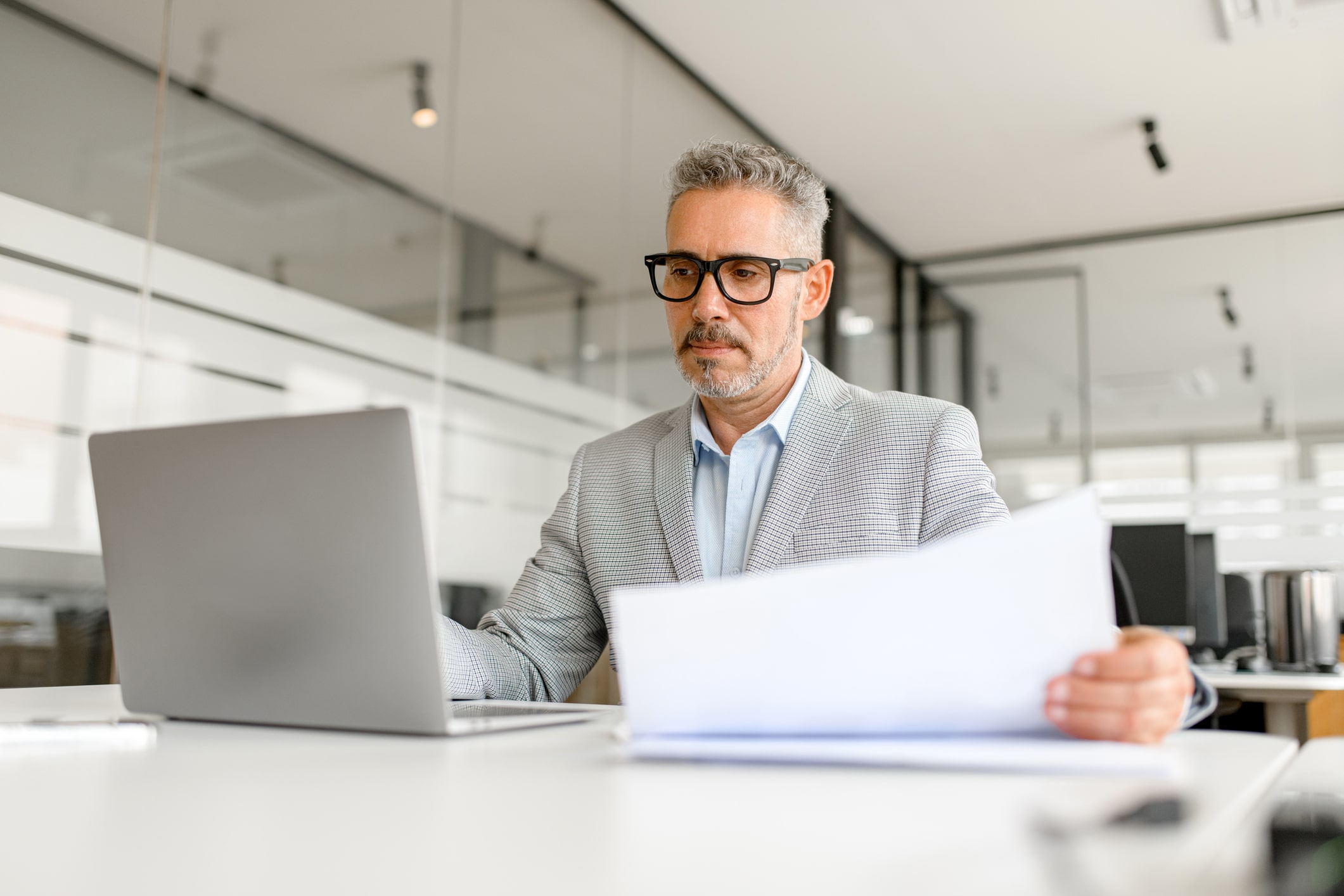 Concentrated middle age male entrepreneur using laptop, confident gray-haired man preparing annual report, working on a new startup sitting at the desk, doing paperwork