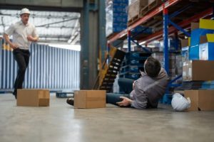 Warehouse Accident, Man worker lies on the ground after a fall while lifting boxes to shelf factory store. Male colleagues rush to assist. Safety, Industry, Healthcare, Insurance Concept.