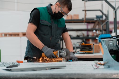 Heavy Industry Engineering Factory Interior with Industrial Worker Using Angle Grinder and Cutting a Metal Tube.He Wears a Mask on His Face Because of the Coronavirus Pandemic. High quality photo