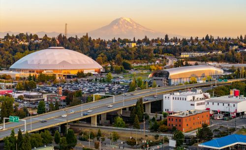 The Tacoma cityscape and its skyscrapers are dramatically framed against the backdrop of the towering, snow-capped Mount Rainier at sunset.
