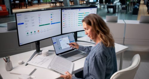 Woman Reviews Payroll Documents While Working On Laptop At Office Desk.