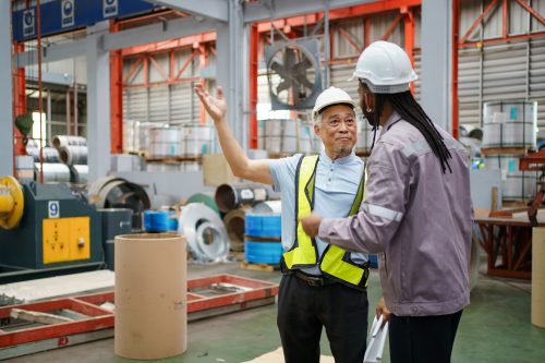 Senior engineer and factory worker discuss about metal production blueprints working together. Teamwork, expertise, and communication in a modern industrial manufacturing environment.