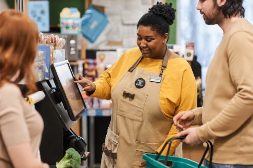 store clerk wearing an apron with a Black Lives Matter button helping someone use self checkout