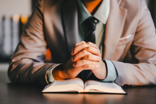 Person in professional clothing with clasped hands praying on an open bible, reflecting devotion, faith, hope, and spirituality, finding comfort in christian religious practice